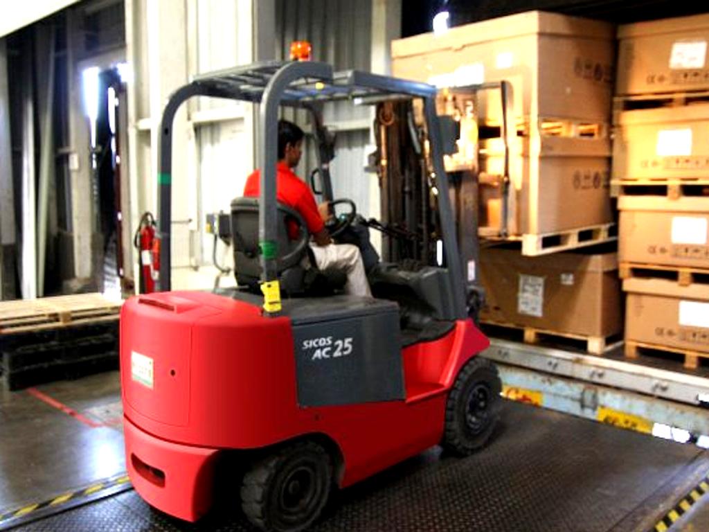 Formation CACES autorisation de conduite : Un cariste opérant un chariot élévateur dans un entrepôt, transportant des boîtes en bois empilées sur des palettes. Le cariste porte un gilet de haute visibilité et se trouve à l'intérieur d'un entrepôt bien éclairé avec des étagères remplies de produits en arrFormation CACES chariot élévateur et chariot automoteur : Cariste en gilet haute visibilité opérant un chariot élévateur, transportant des Palettes dans un entrepôt. La formation autorisation de conduite inclut l'utilisation de chariots élévateurs et automoteurs pour une meilleure sécurité et efficacité en milieu logistique.